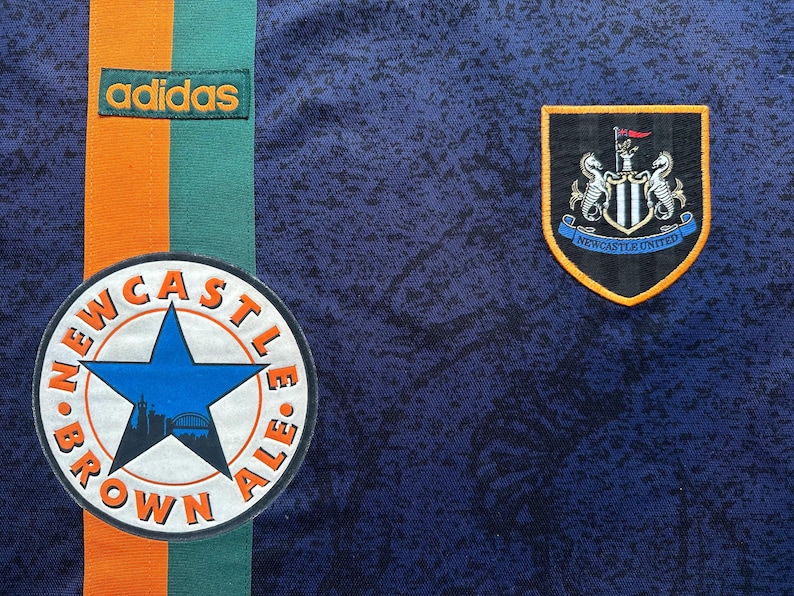 May include: Close-up of a navy blue soccer jersey with orange and green stripes. Features the Adidas logo, a Newcastle United crest, and a Newcastle Brown Ale patch.