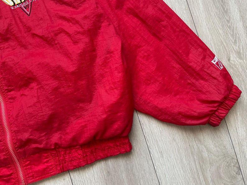 May include: A bright red vintage jacket with a zipper closure and elastic cuffs. The jacket has a logo on the upper left chest and a small logo on the sleeve. The fabric has a slight sheen.