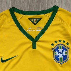 Pu&ograve; includere: Primo piano di una maglia da calcio brasiliana gialla con finiture verdi. Lo scollo a V e le maniche sono rifiniti in verde. Il logo Nike e lo stemma CBF con cinque stelle sono visibili. La maglia &egrave; realizzata in tessuto strutturato.