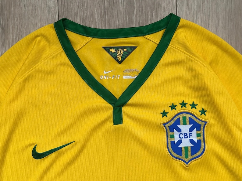 Pu&ograve; includere: Primo piano di una maglia da calcio brasiliana gialla con finiture verdi. Lo scollo a V e le maniche sono rifiniti in verde. Il logo Nike e lo stemma CBF con cinque stelle sono visibili. La maglia &egrave; realizzata in tessuto strutturato.