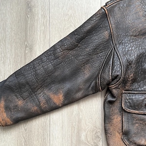 May include: A dark brown leather jacket featuring a heavily distressed, textured finish with lighter tan worn areas on the sleeve and body. The jacket includes a ribbed cuff, a structured shoulder seam, and a front pocket with a single brown button.