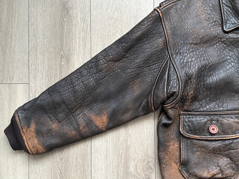 May include: A dark brown leather jacket featuring a heavily distressed, textured finish with lighter tan worn areas on the sleeve and body. The jacket includes a ribbed cuff, a structured shoulder seam, and a front pocket with a single brown button.
