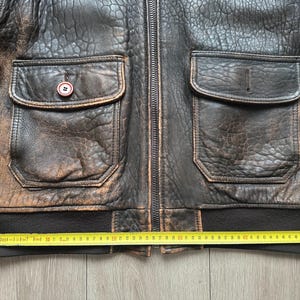 May include: A dark brown distressed leather bomber jacket with a central zip, two large front flap pockets, and a ribbed hem. A yellow measuring tape is positioned across the bottom, showing a width of approximately 55 centimetres.