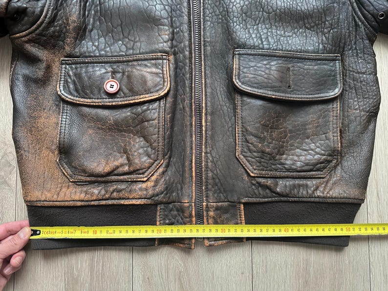 May include: A dark brown distressed leather bomber jacket with a central zip, two large front flap pockets, and a ribbed hem. A yellow measuring tape is positioned across the bottom, showing a width of approximately 55 centimetres.