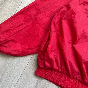 May include: Close-up of a bright red jacket. The jacket features a gathered elastic waistband and cuffs, and a side pocket. The fabric has a slight sheen, suggesting a nylon or similar material. The jacket's vibrant color is eye-catching.
