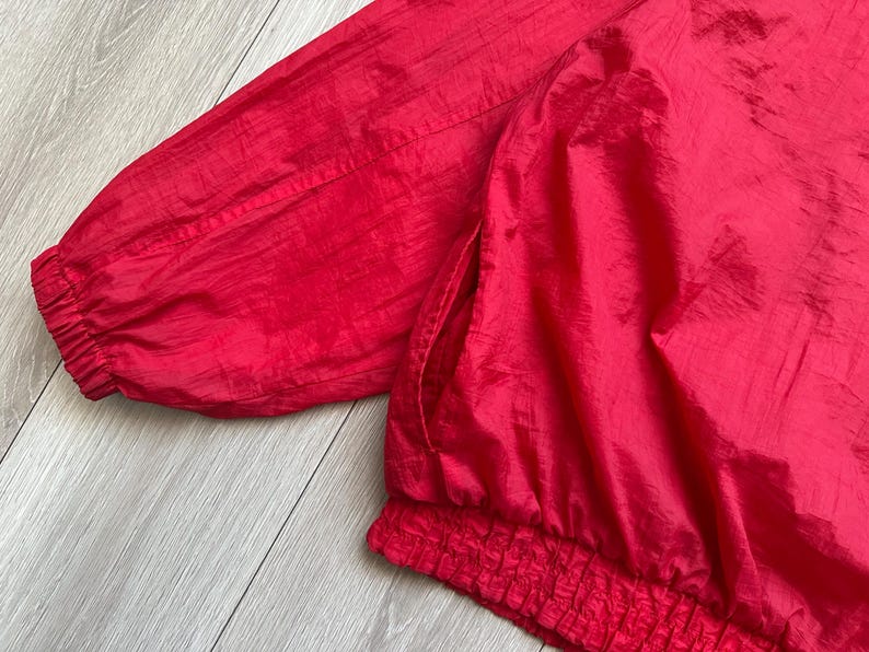 May include: Close-up of a bright red jacket. The jacket features a gathered elastic waistband and cuffs, and a side pocket. The fabric has a slight sheen, suggesting a nylon or similar material. The jacket's vibrant color is eye-catching.