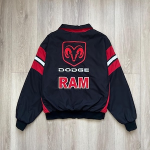 Puede incluir: Chaqueta negra con detalles rojos y blancos. La parte trasera presenta un logotipo rojo de Dodge Ram y las palabras "DODGE RAM" en blanco. Tiene cuello y puños acanalados.