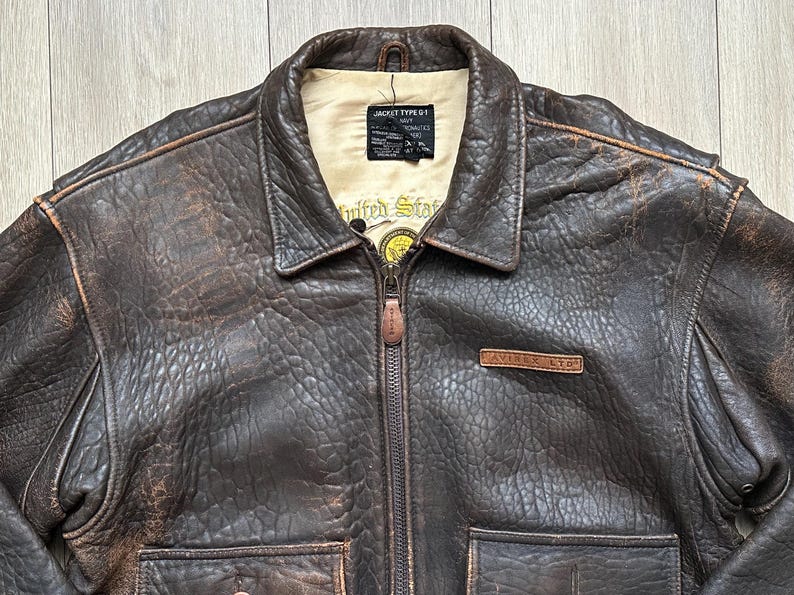 May include: A dark brown distressed leather bomber jacket with a cream-coloured interior lining. The jacket features a front zip closure, a fold-down collar, two front pockets, and a leather patch on the chest reading AVIREX LTD. A black label inside reads JACKET TYPE G-1.