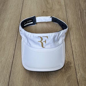 Può includere: Visiera bianca con le iniziali dorate "RF" ricamate sul davanti. La visiera ha una fascia interna nera e un piccolo logo Nike. Realizzata in materiale leggero e traspirante, è ideale per lo sport e le attività all'aperto.