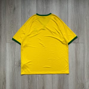 Pu&ograve; includere: Una maglietta gialla con rifiniture verdi intorno al colletto e alle maniche. La maglietta &egrave; realizzata in un materiale morbido ed &egrave; stesa su una superficie di legno. La maglietta &egrave; di colore giallo pieno.