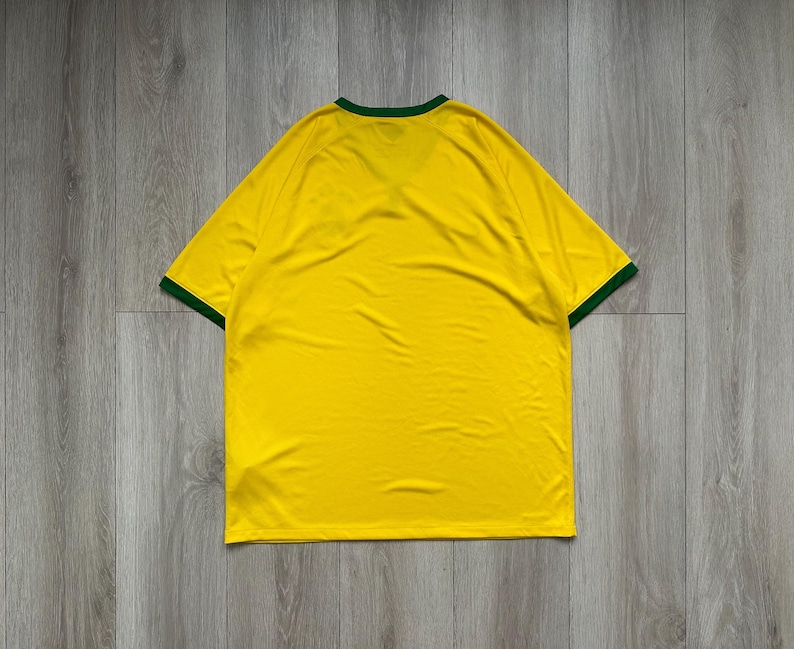 Pu&ograve; includere: Una maglietta gialla con rifiniture verdi intorno al colletto e alle maniche. La maglietta &egrave; realizzata in un materiale morbido ed &egrave; stesa su una superficie di legno. La maglietta &egrave; di colore giallo pieno.