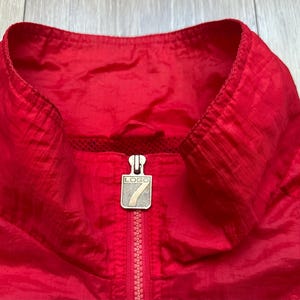May include: Close-up of a bright red jacket collar with a zipper. The jacket has a high collar and a silver zipper pull with the word "LOGO" on it. The material appears to be a lightweight, possibly nylon, fabric.