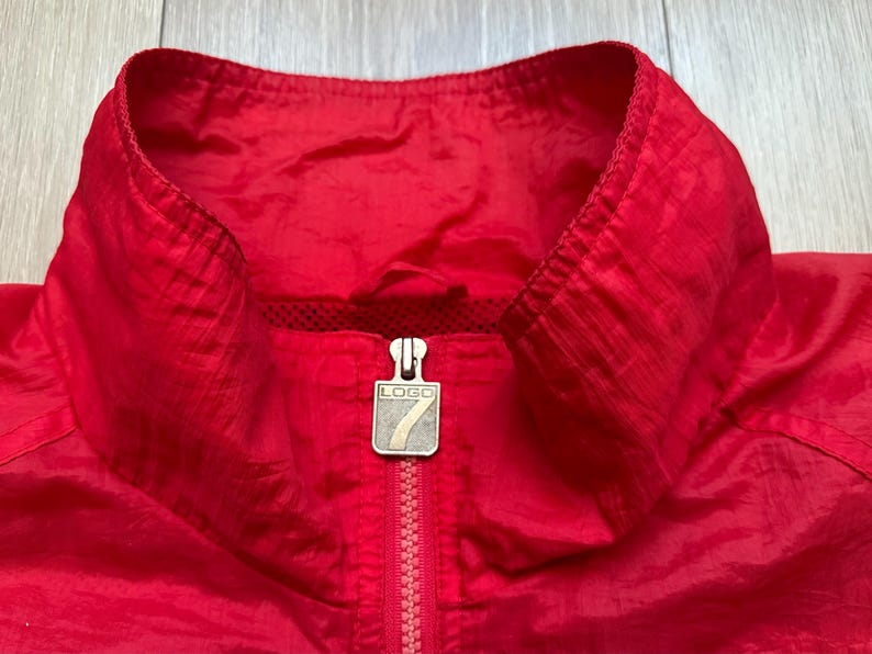 May include: Close-up of a bright red jacket collar with a zipper. The jacket has a high collar and a silver zipper pull with the word "LOGO" on it. The material appears to be a lightweight, possibly nylon, fabric.