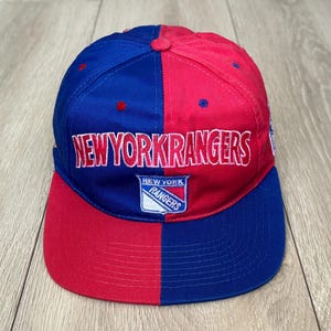 May include: A split-colour baseball cap featuring a blue left side and a red right side. The front displays the text NEW YORK RANGERS in white embroidery with red outlines, positioned above a New York Rangers team logo patch in blue, red, and white.