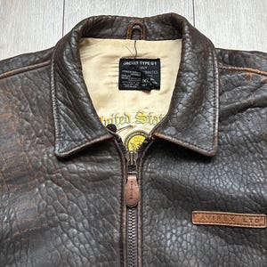 May include: A dark brown distressed leather jacket with a pointed collar and a front zip fastening. The interior features a cream-coloured lining with a black label reading JACKET TYPE G-1 and a yellow circular emblem with the text United States.