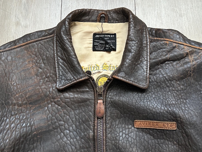 May include: A dark brown distressed leather jacket with a pointed collar and a front zip fastening. The interior features a cream-coloured lining with a black label reading JACKET TYPE G-1 and a yellow circular emblem with the text United States.