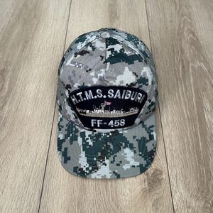May include: A grey and green camouflage baseball cap. The cap has a patch with the text "H.T.M.S. SAIBURI" above an image of a ship and the text "FF-458". The cap has a curved peak.