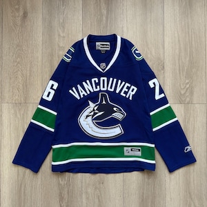 May include: A blue Vancouver Canucks hockey jersey with white and green accents. The jersey features the team's logo, the word "VANCOUVER", and the numbers 6 and 2. The Reebok logo is visible.