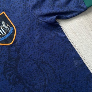 May include: A navy blue Newcastle United soccer jersey with a black and blue pattern. The jersey features the team's crest on the chest, with a black, white, and orange design. The sleeve cuff is green.