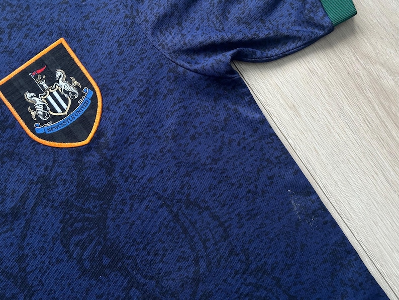 May include: A navy blue Newcastle United soccer jersey with a black and blue pattern. The jersey features the team's crest on the chest, with a black, white, and orange design. The sleeve cuff is green.
