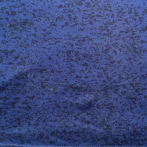 May include: Close-up of a blue fabric with a speckled pattern. The fabric has a textured appearance, with small, dark blue shapes scattered across the surface. The overall effect is a mottled design, creating visual interest.