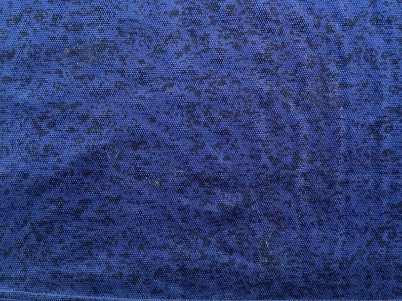 May include: Close-up of a blue fabric with a speckled pattern. The fabric has a textured appearance, with small, dark blue shapes scattered across the surface. The overall effect is a mottled design, creating visual interest.