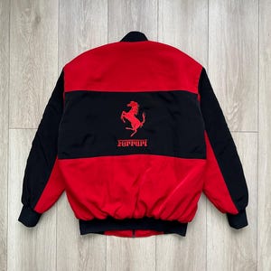 May include: A red and black bomber jacket with the Ferrari logo. The jacket features a red body with black sleeves and a black horizontal stripe across the chest. The Ferrari logo is in red.