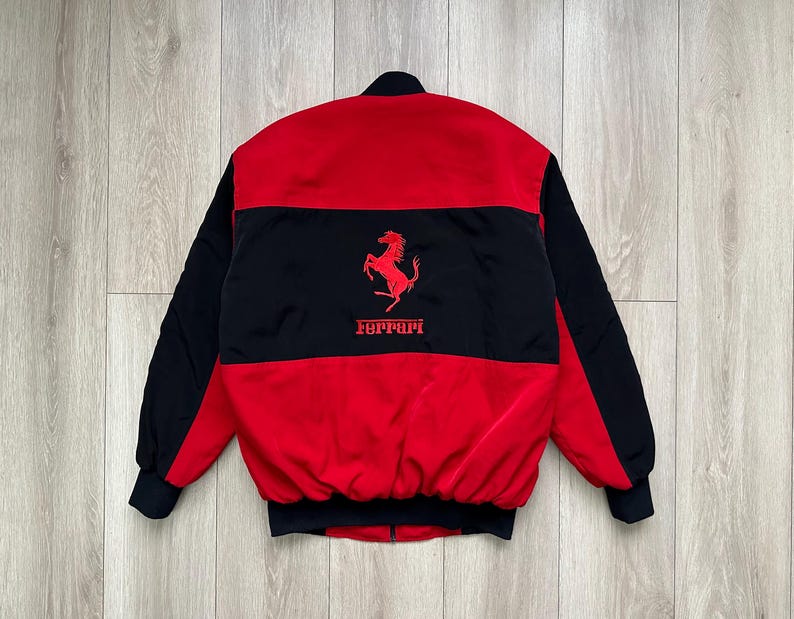 May include: A red and black bomber jacket with the Ferrari logo. The jacket features a red body with black sleeves and a black horizontal stripe across the chest. The Ferrari logo is in red.