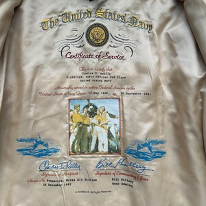 May include: A cream-coloured jacket lining featuring an embroidered United States Navy Certificate of Service for Charles T. Reilly. The text includes dates, rank, and signatures, with a central colour photograph of sailors and blue nautical graphics.