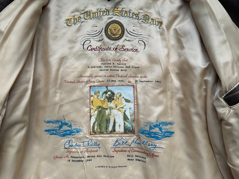 May include: A cream-coloured jacket lining featuring an embroidered United States Navy Certificate of Service for Charles T. Reilly. The text includes dates, rank, and signatures, with a central colour photograph of sailors and blue nautical graphics.