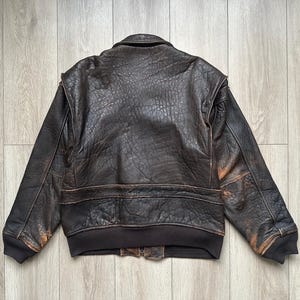 May include: A dark brown leather bomber jacket with a distressed, textured finish. The jacket features a fold-down collar, long sleeves with ribbed cuffs, and a ribbed hem. The leather shows signs of wear with lighter brown highlights.
