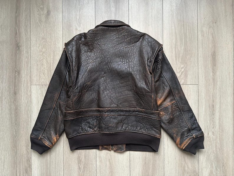 May include: A dark brown leather bomber jacket with a distressed, textured finish. The jacket features a fold-down collar, long sleeves with ribbed cuffs, and a ribbed hem. The leather shows signs of wear with lighter brown highlights.