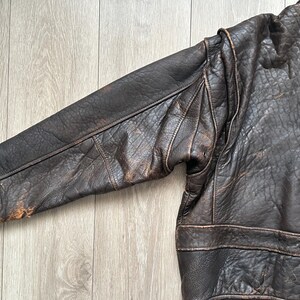 May include: A dark brown leather bomber jacket featuring a distressed, textured finish, long sleeves with reinforced seams, and a dark brown ribbed knit cuff at the wrist. The jacket displays visible signs of wear and natural leather grain patterns.
