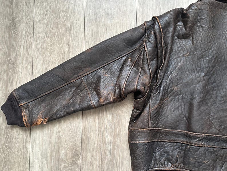 May include: A dark brown leather bomber jacket featuring a distressed, textured finish, long sleeves with reinforced seams, and a dark brown ribbed knit cuff at the wrist. The jacket displays visible signs of wear and natural leather grain patterns.