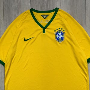 Pu&ograve; includere: Maglia da calcio gialla del Brasile con finiture verdi su colletto e maniche. Presenta il logo Nike verde e uno stemma CBF con cinque stelle. La maglia ha uno scollo a V.