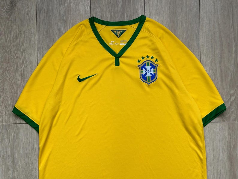 Pu&ograve; includere: Maglia da calcio gialla del Brasile con finiture verdi su colletto e maniche. Presenta il logo Nike verde e uno stemma CBF con cinque stelle. La maglia ha uno scollo a V.