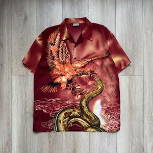May include: A short-sleeve button-down shirt in deep red with a graphic print of an eagle descending toward a coiled snake. The shirt features a camp collar, a button-front closure, and a vibrant, fiery-toned illustration across the front panel.