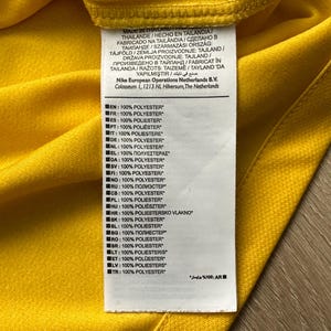 Pu&ograve; includere: Primo piano di un capo giallo con un'etichetta bianca. L'etichetta mostra testo in pi&ugrave; lingue, tra cui inglese, francese e spagnolo, che indica che l'articolo &egrave; prodotto in Thailandia ed &egrave; 100% poliestere. &Egrave; visibile anche il logo Nike.
