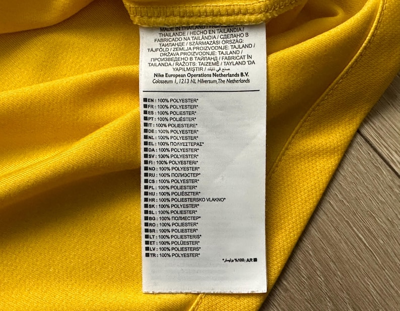 Pu&ograve; includere: Primo piano di un capo giallo con un'etichetta bianca. L'etichetta mostra testo in pi&ugrave; lingue, tra cui inglese, francese e spagnolo, che indica che l'articolo &egrave; prodotto in Thailandia ed &egrave; 100% poliestere. &Egrave; visibile anche il logo Nike.