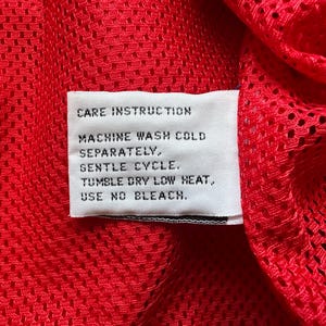 May include: Close-up of a red mesh fabric with a white care instruction label. The label reads: Machine wash cold separately, gentle cycle, tumble dry low heat, use no bleach.