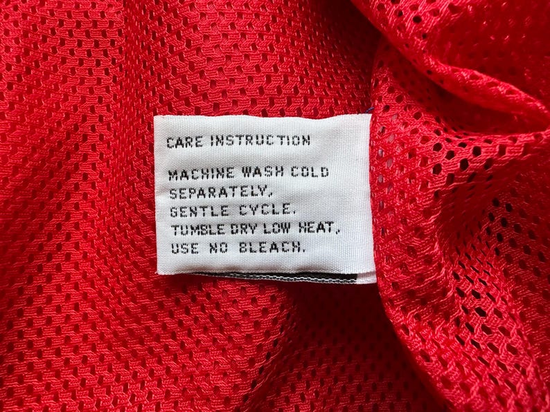 May include: Close-up of a red mesh fabric with a white care instruction label. The label reads: Machine wash cold separately, gentle cycle, tumble dry low heat, use no bleach.