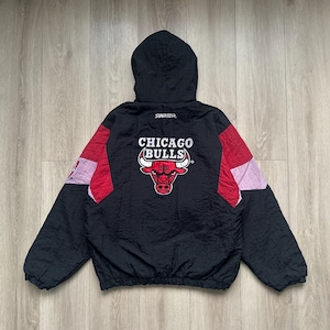 Puede incluir: Chaqueta negra de los Chicago Bulls con capucha. La chaqueta presenta paneles rojos y rosas en las mangas y el logotipo de los Chicago Bulls en la espalda. El logotipo de Starter es visible en la capucha.