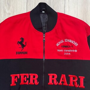May include: Red and black Ferrari Formula 1 jacket. The jacket features the Ferrari logo, the words "FER RARI" in large red letters, and the text "MICHAEL SCHUMACHER FORMULA-1 World championship 2004". The jacket has a black collar and a zipper.