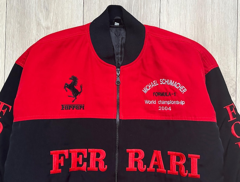 May include: Red and black Ferrari Formula 1 jacket. The jacket features the Ferrari logo, the words "FER RARI" in large red letters, and the text "MICHAEL SCHUMACHER FORMULA-1 World championship 2004". The jacket has a black collar and a zipper.