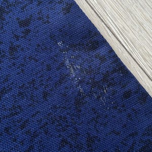 May include: Close-up of a section of dark blue fabric with a black pattern. The fabric has a textured appearance. A small area of white discoloration is visible on the fabric. The fabric is next to a light-colored wooden surface.