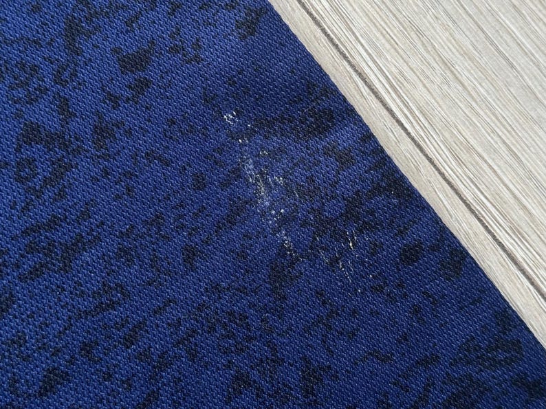 May include: Close-up of a section of dark blue fabric with a black pattern. The fabric has a textured appearance. A small area of white discoloration is visible on the fabric. The fabric is next to a light-colored wooden surface.