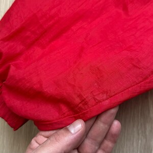 May include: Close-up of a bright red garment, possibly a jacket or windbreaker, with a gathered elastic cuff. The fabric has a slight sheen, suggesting a synthetic material. The texture is smooth and slightly wrinkled.