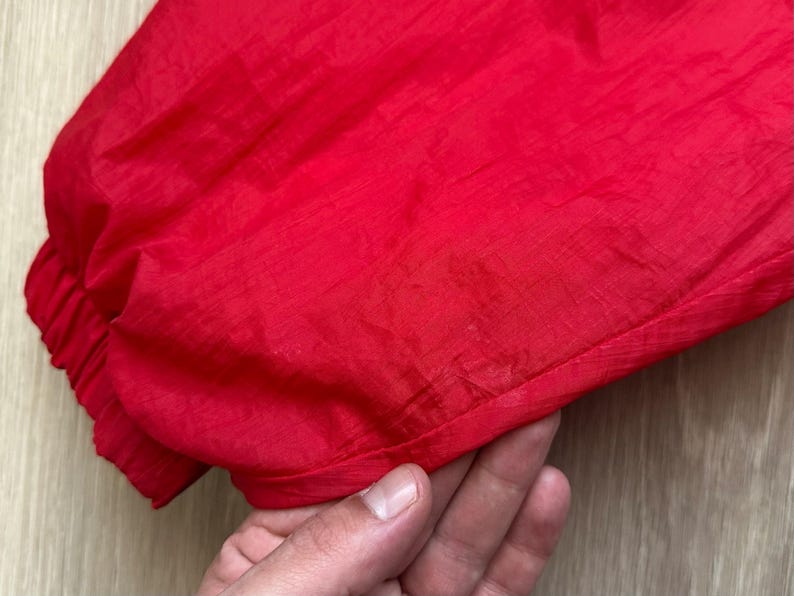 May include: Close-up of a bright red garment, possibly a jacket or windbreaker, with a gathered elastic cuff. The fabric has a slight sheen, suggesting a synthetic material. The texture is smooth and slightly wrinkled.