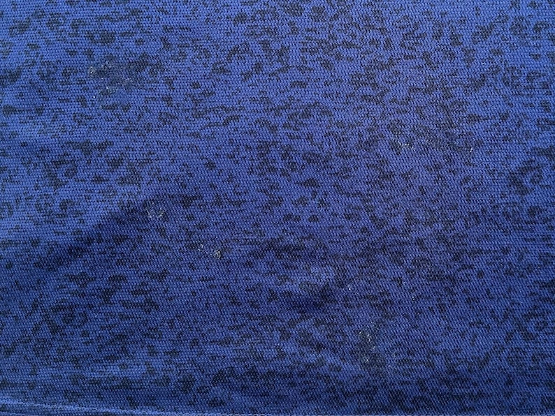 May include: Close-up of a blue fabric with a textured surface. The fabric has a pattern of dark blue, irregular shapes scattered across its surface. The overall appearance is a mottled or speckled design.