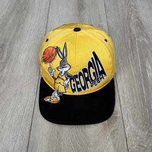 May include: Yellow and black baseball cap featuring Bugs Bunny in a basketball uniform. The cap has the words "GEORGIA TECH" in black lettering. The brim is black, and the top is yellow.